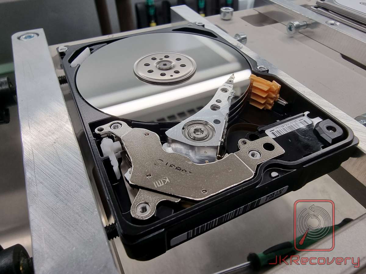 Otwarty dysk twardy HDD z widocznym talerzem magnetycznym, ramieniem głowicy i mechanizmem napędowym, umieszczony w uchwycie laboratoryjnym podczas prac serwisowych
