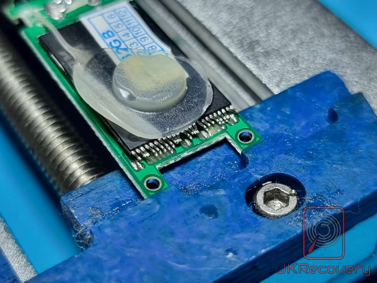 Mała płytka PCB z układem scalonym pokrytym przezroczystą żywicą epoksydową, umieszczona w niebieskim imadle warsztatowym podczas procesu odzysku danych