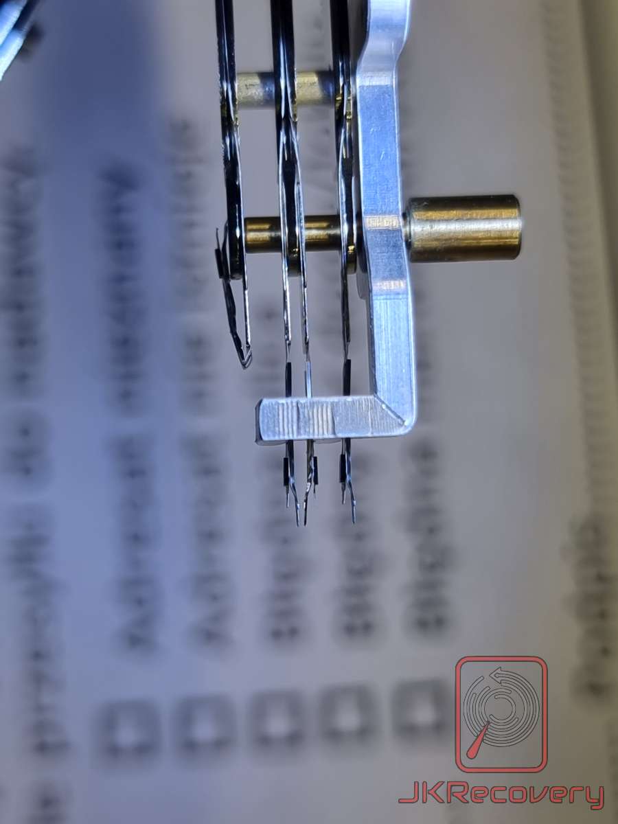 Zbliżenie na głowice odczytu/zapisu dysku twardego HDD zamontowane w specjalnym uchwycie serwisowym z mosiężnymi elementami dystansowymi, widoczne na tle dokumentacji technicznej