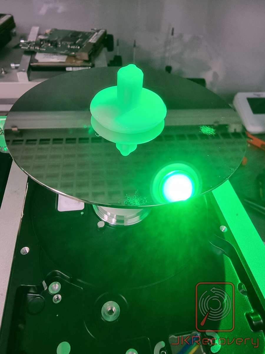 Otwarty dysk twardy HDD z widocznym talerzem magnetycznym oświetlonym zielonym światłem UV w laboratorium odzysku danych JKRecovery