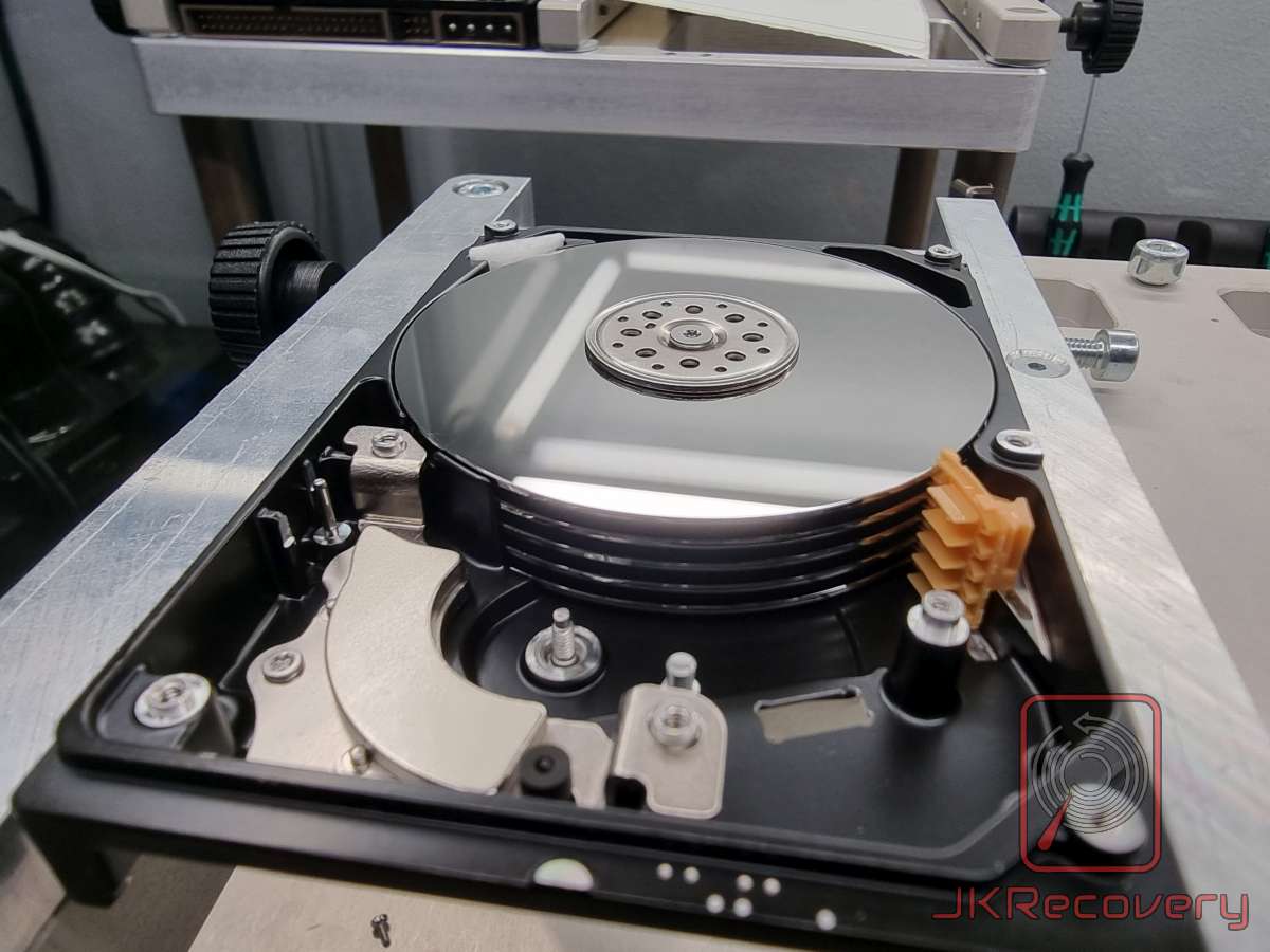 Otwarty dysk twardy HDD zamontowany w specjalistycznym uchwycie laboratoryjnym, widoczne talerze magnetyczne, głowice odczytu/zapisu oraz pomarańczowy plastikowy element dystansowy między talerzami