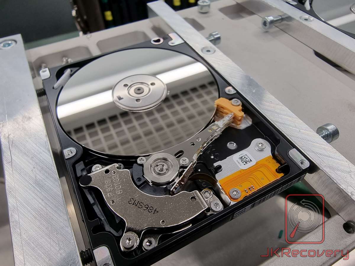 Otwarty dysk twardy HDD zamontowany w metalowym uchwycie w laboratorium odzysku danych, widoczny talerz magnetyczny, głowica odczytu/zapisu oraz ramię aktuatora z pomarańczowym elementem mocującym