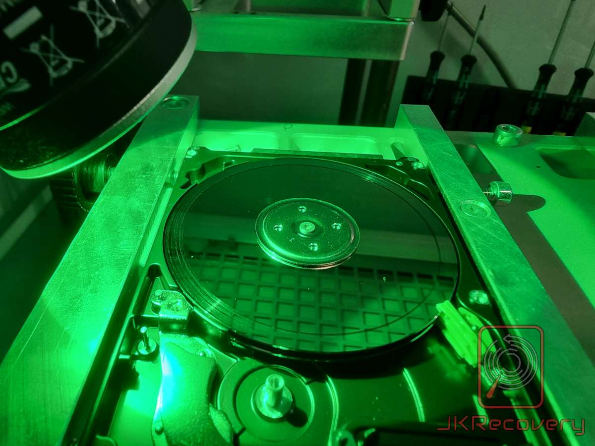 Otwarty dysk twardy HDD umieszczony w specjalistycznym uchwycie, oświetlony zielonym światłem UV w warunkach laboratoryjnych - widoczny talerz magnetyczny i głowice odczytu/zapisu