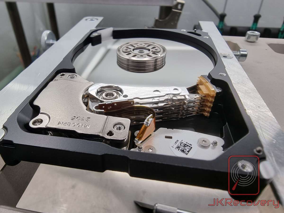 Otwarty dysk twardy HDD umieszczony w specjalnym uchwycie laboratoryjnym - widoczny talerz magnetyczny, wielowarstwowy zespół głowic odczytu/zapisu oraz mechanizm aktuatora z cewką głosową