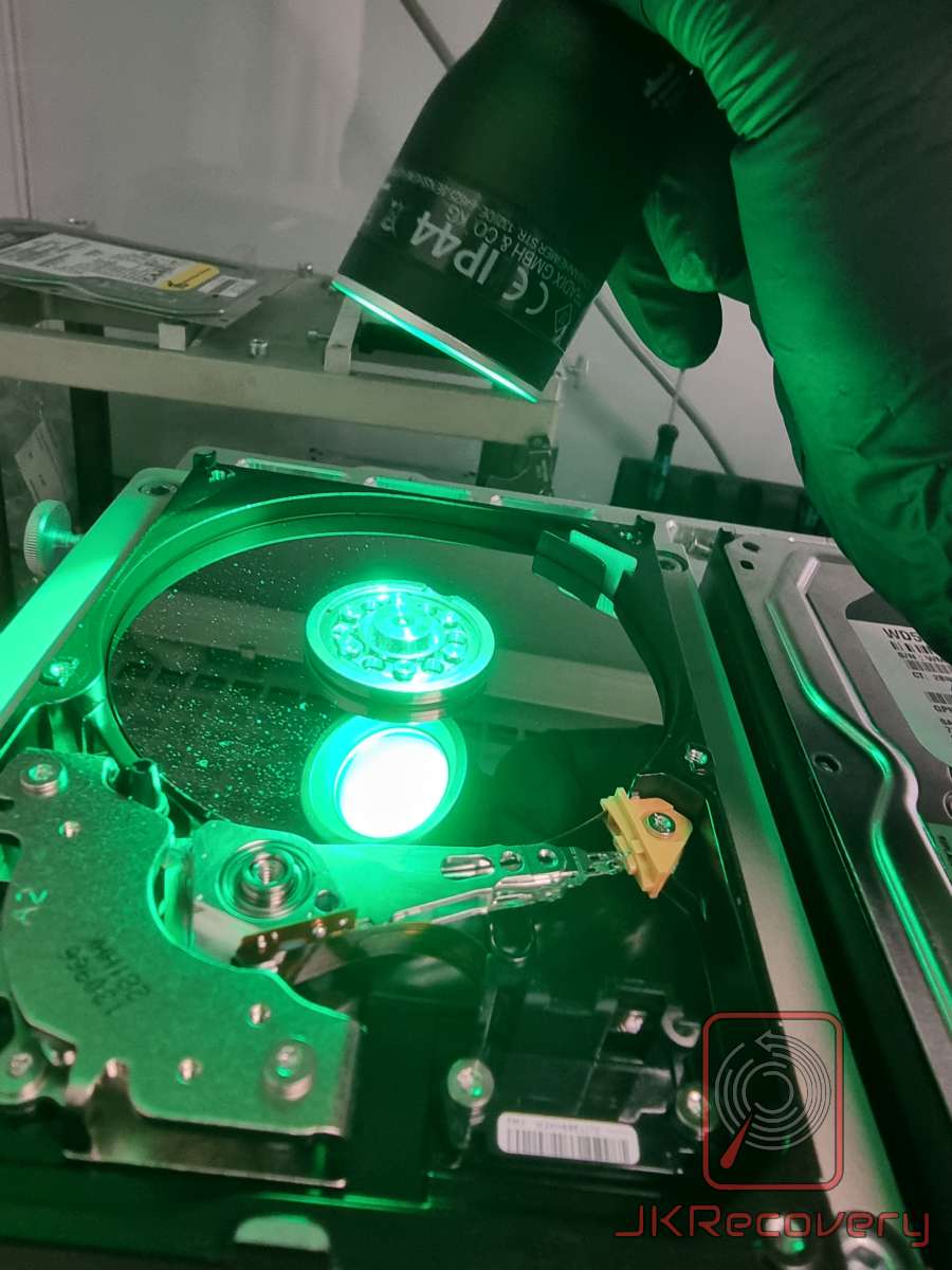 Technik w zielonych rękawiczkach trzymający lampę UV nad otwartym dyskiem twardym HDD, oświetlając talerze dysku zielonym światłem w celu wykrycia zanieczyszczeń lub uszkodzeń powierzchni