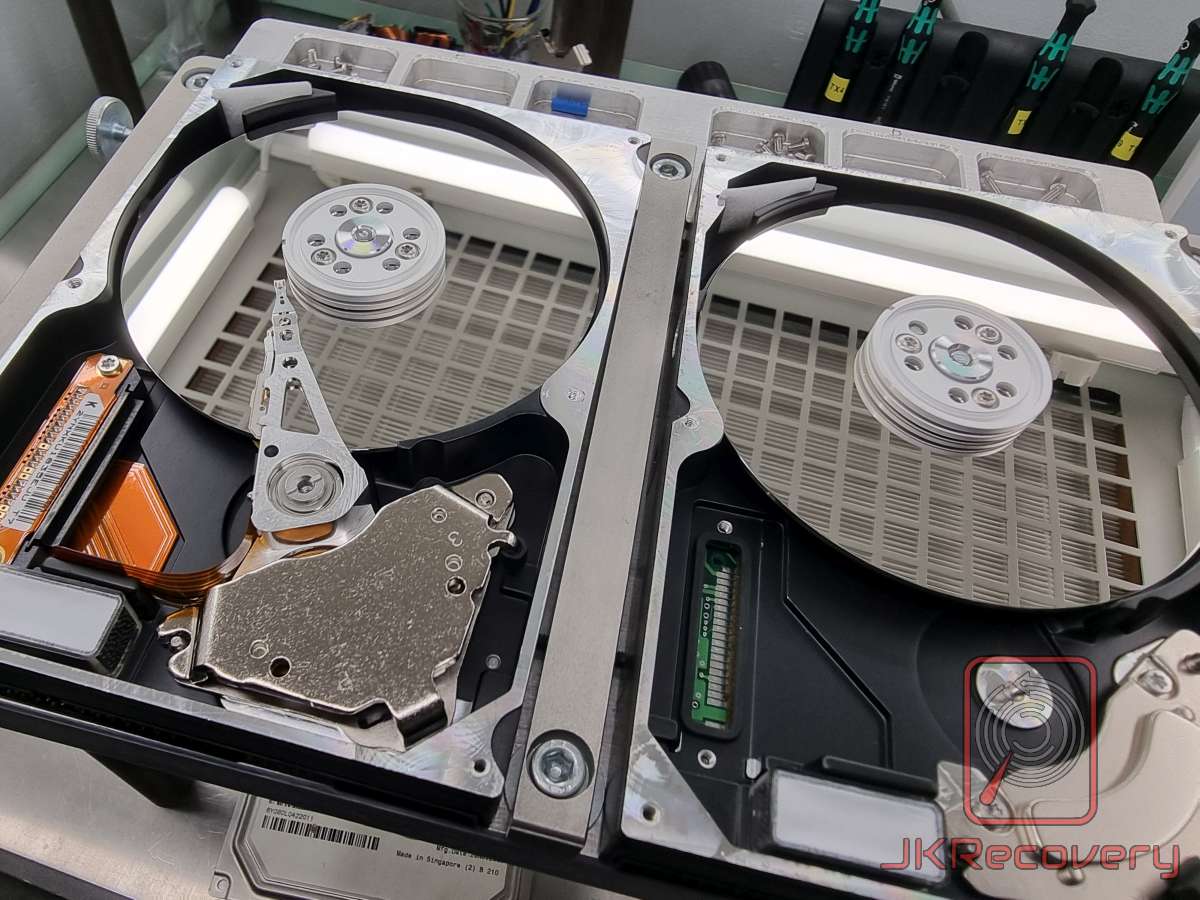 Dwa otwarte dyski twarde HDD umieszczone obok siebie w komorze czystego powietrza. Lewy dysk z widocznym uszkodzonym lub wymienianym ramieniem odczytu/zapisu, prawy dysk jako donor części. Procedura transplantacji głowic w laboratorium odzysku danych JKRecovery.