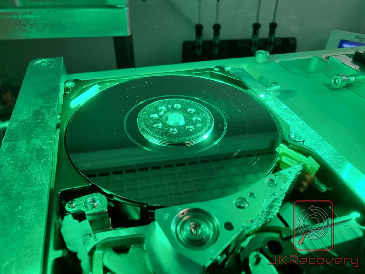 Widok z góry na otwarty dysk twardy HDD zamontowany w uchwycie laboratoryjnym, oświetlony zielonym światłem, widoczny talerz magnetyczny z centralnym mocowaniem i śrubami oraz fragment mechanizmu ramienia głowicy