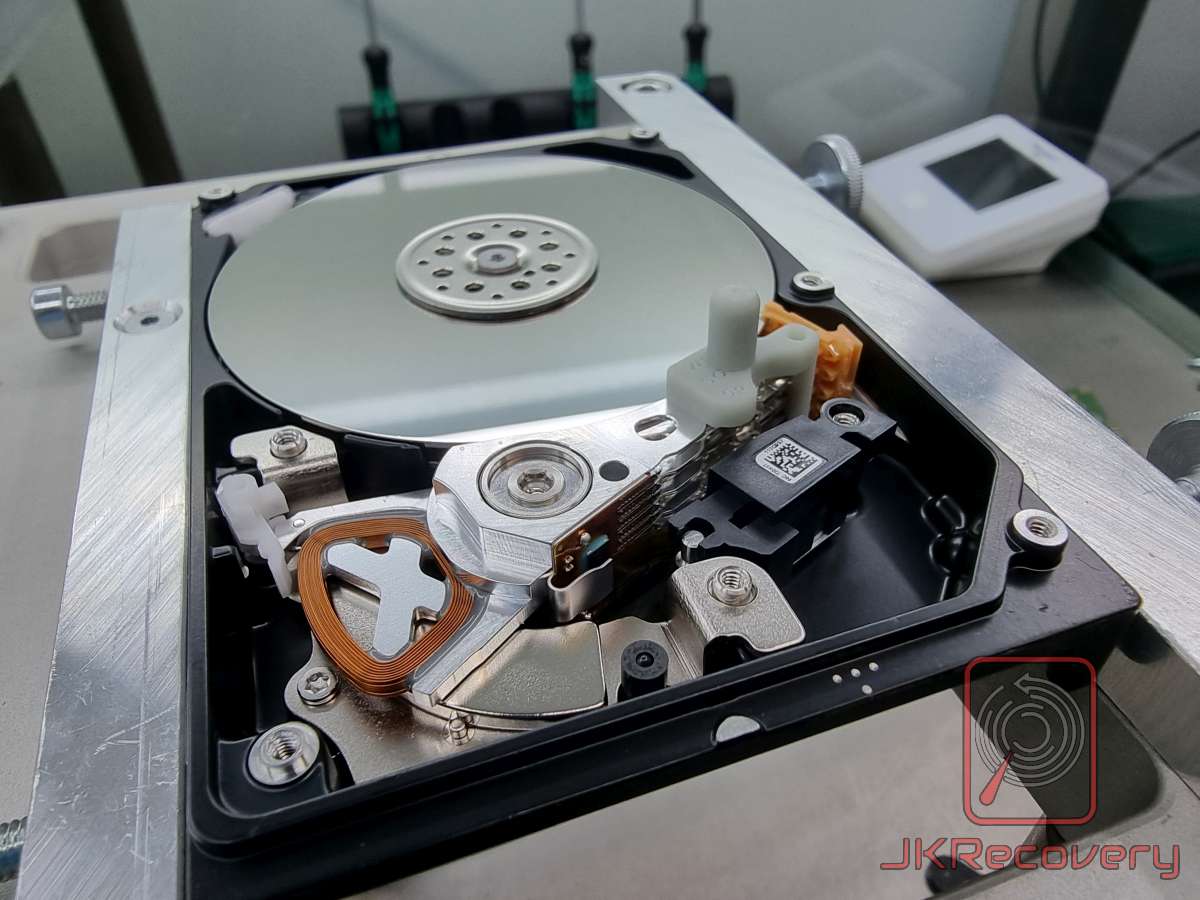 Otwarty dysk twardy HDD w laboratorium odzysku danych JKRecovery – widoczne ramię głowicy odczytu/zapisu, cewka VCM, talerz magnetyczny oraz mechanizm parkowania głowicy, dysk umieszczony w profesjonalnym uchwycie serwisowym
