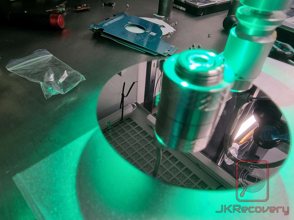 Talerz dysku twardego HDD zamontowany na specjalistycznym uchwycie w laboratorium odzysku danych JKRecovery, w tle uszkodzona płyta PCB dysku i woreczek strunowy z podzespołami