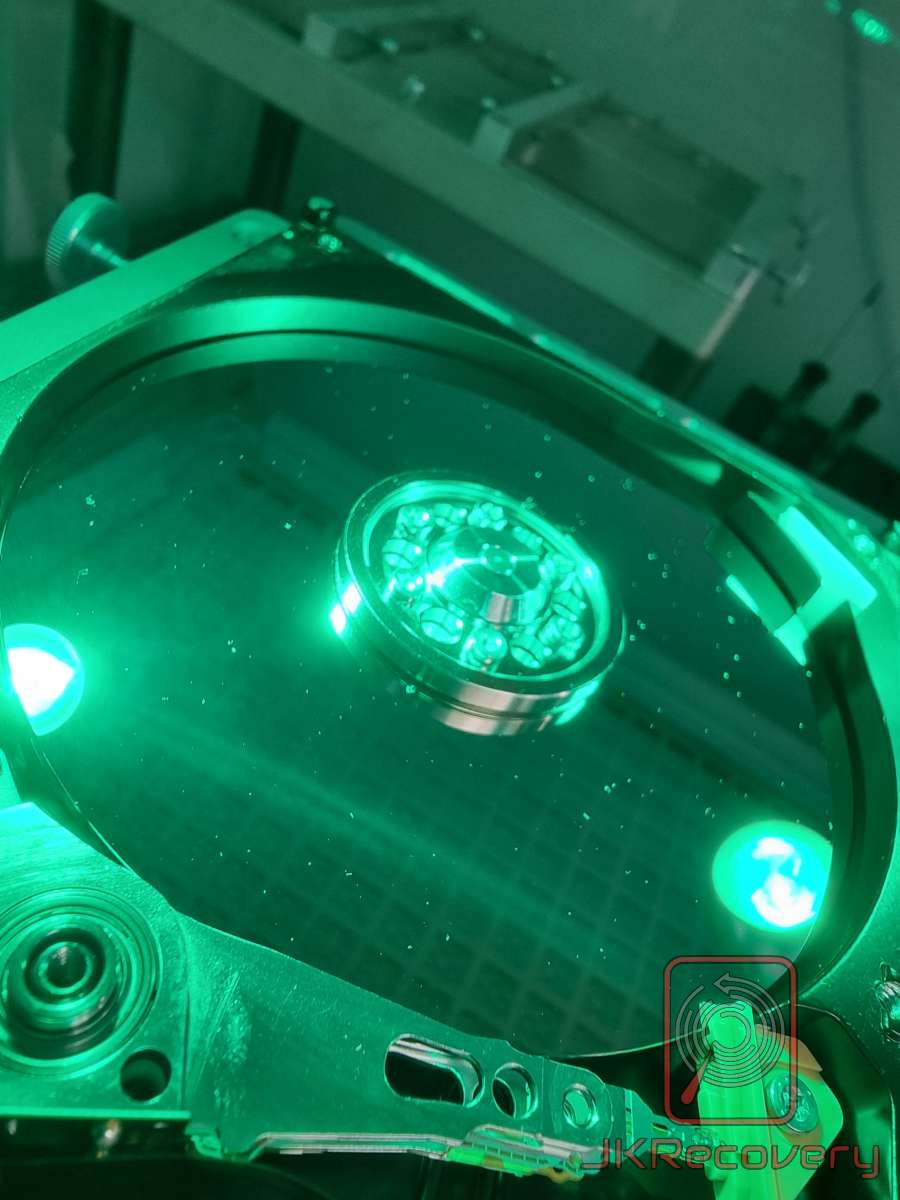 Wnętrze otwartego dysku twardego HDD oświetlone zielonym światłem UV, widok na talerz magnetyczny i obudowę dysku podczas diagnostyki w laboratorium odzysku danych JKRecovery