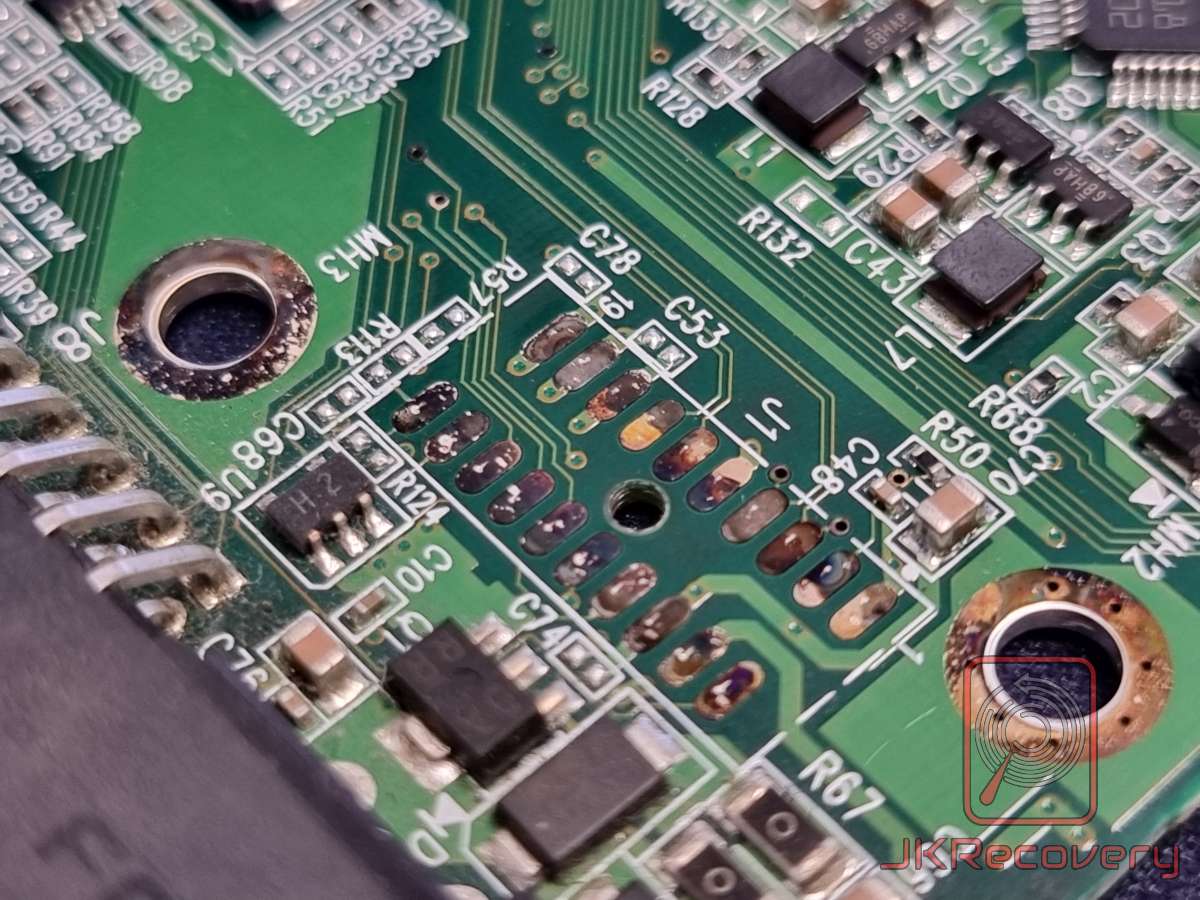 Zbliżenie na uszkodzoną płytę PCB dysku twardego HDD z widocznymi śladami korozji, przepalenia i uszkodzeń komponentów SMD w obszarze kondensatorów i rezystorów