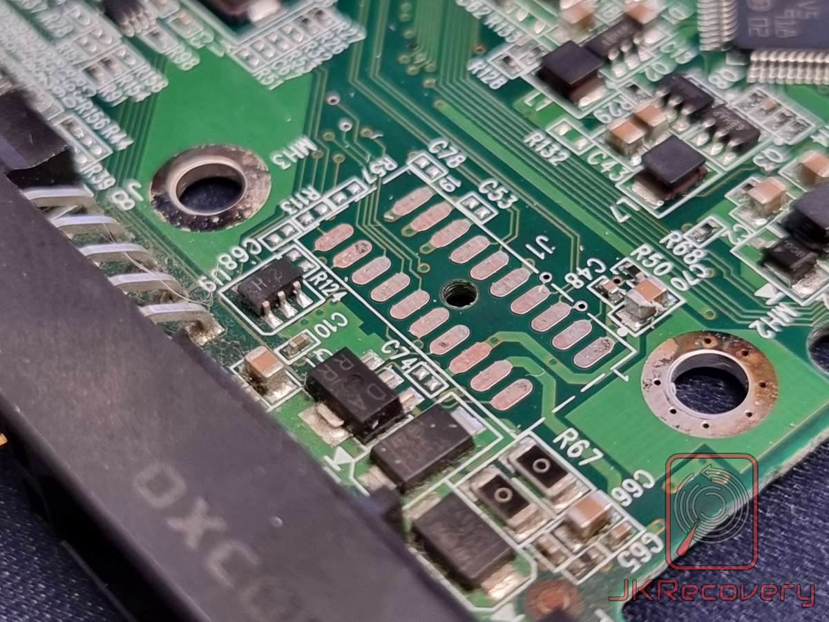 Zbliżenie na płytę PCB dysku twardego HDD - widoczne komponenty elektroniczne, rezystory SMD, kondensatory, diody oraz złącze J1 z pustymi padami lutowniczymi na zielonej płytce drukowanej