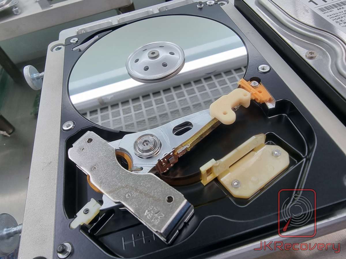 Otwarty dysk twardy HDD w laboratorium czystym JKRecovery - widoczny talerz magnetyczny, głowica odczytu/zapisu oraz ramię aktuatora z pomarańczowym i beżowym mechanizmem blokady