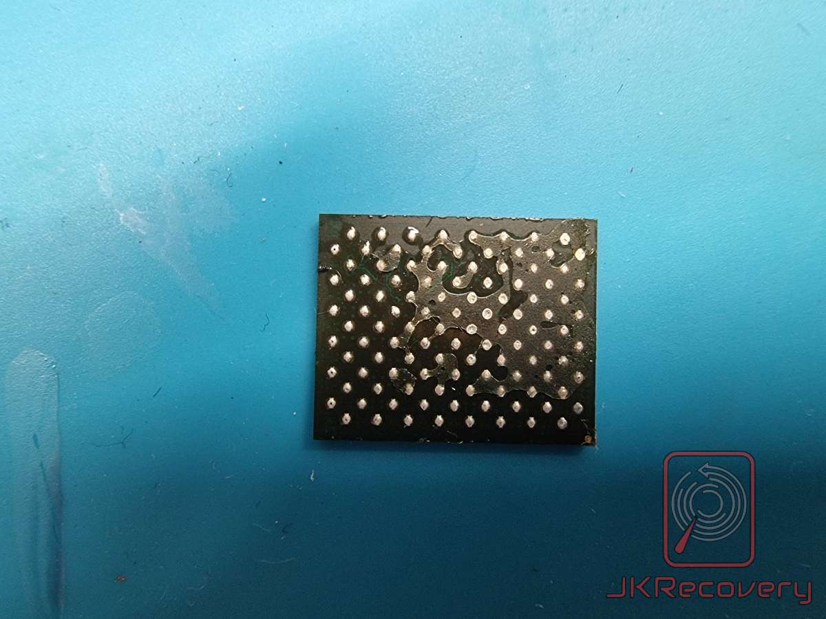 Układ scalony BGA z widocznymi kulkami lutowniczymi, zdjęty z płytki PCB, leżący na niebieskiej podkładce antystatycznej. Widoczne uszkodzenia powłoki ochronnej układu.