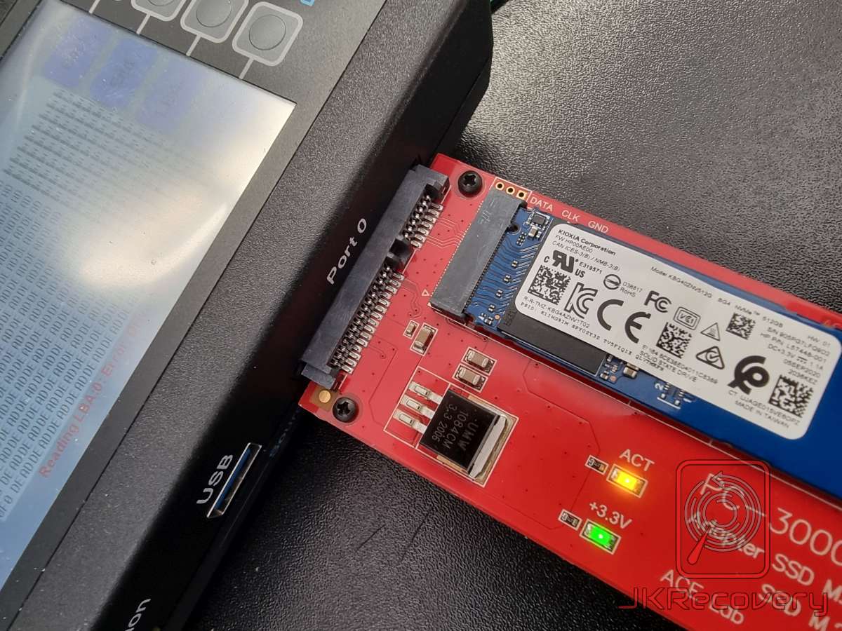 Dysk NVMe Kioxia 512GB podłączony do czytnika PC-3000 SSD M.2 w kolorze czerwono-niebieskim, widoczne diody ACT i +3.3V oraz ekran urządzenia z komunikatem odczytu w tle