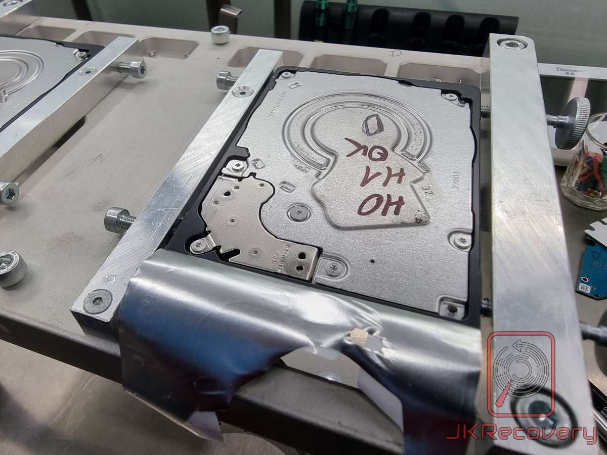 Dysk twardy HDD zamocowany w aluminiowym uchwycie laboratoryjnym, na obudowie napis markerem 'HO HV OK', dysk zabezpieczony czarną taśmą, widok stanowiska roboczego laboratorium odzysku danych JKRecovery
