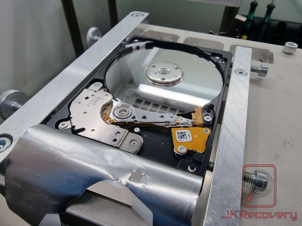 Otwarty dysk twardy HDD zamontowany w specjalistycznym uchwycie laboratoryjnym, widoczne talerze magnetyczne, ramię odczytu/zapisu z głowicami oraz mechanizm aktuatora