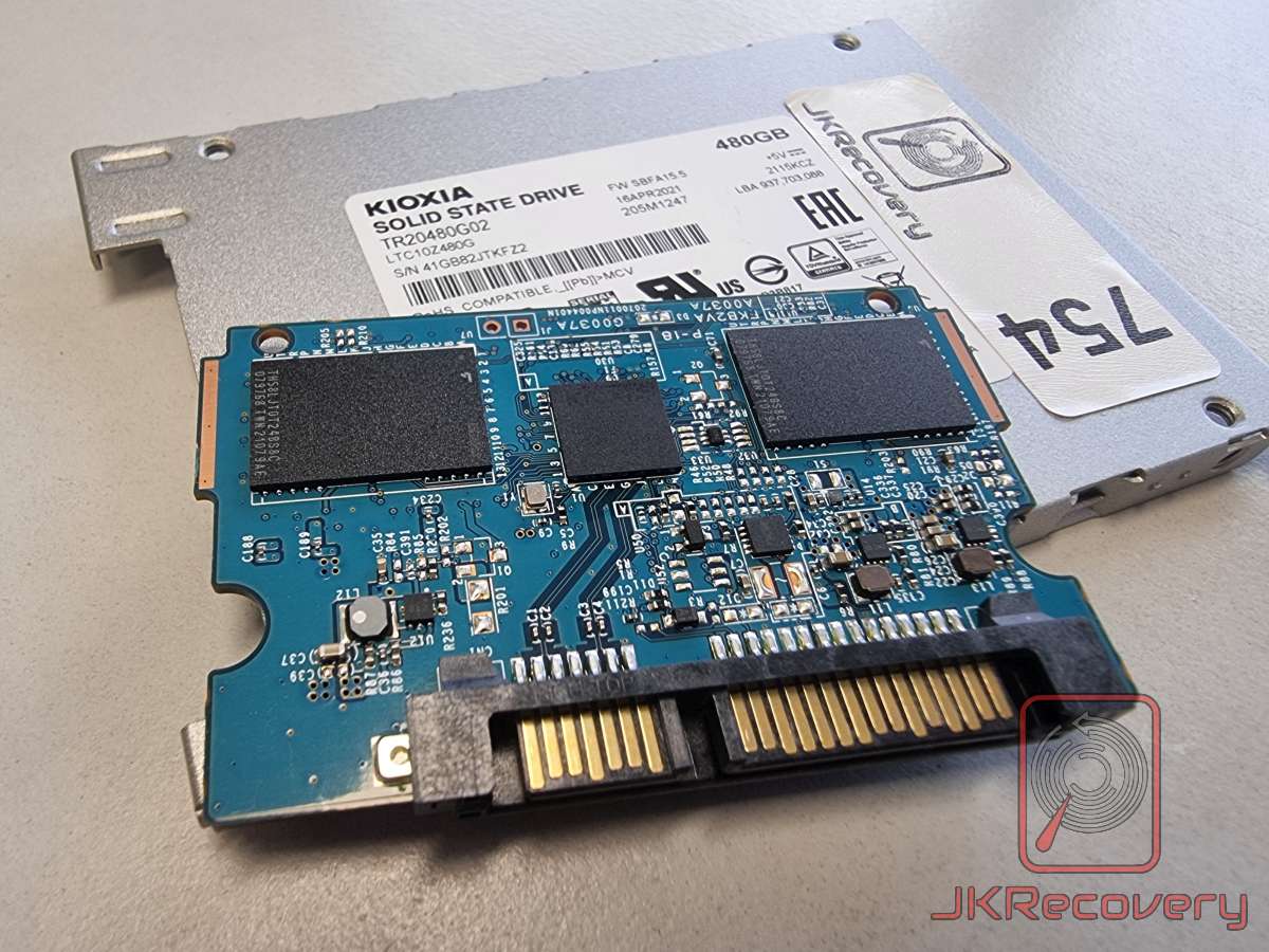 Rozmontowany dysk SSD Kioxia TR20480G02 480GB z widoczną płytą PCB w kolorze niebieskim, złączem SATA oraz układami pamięci flash. W tle widoczna metalowa obudowa z naklejką znamionową.