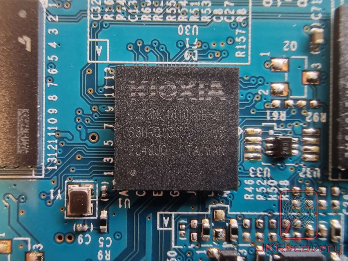 Zbliżenie na kontroler lub układ pamięci NAND flash firmy Kioxia na niebieskiej płycie PCB dysku SSD. Na układzie widoczne oznaczenie TC58NC1010GSE oraz kod produkcji 2049UO.