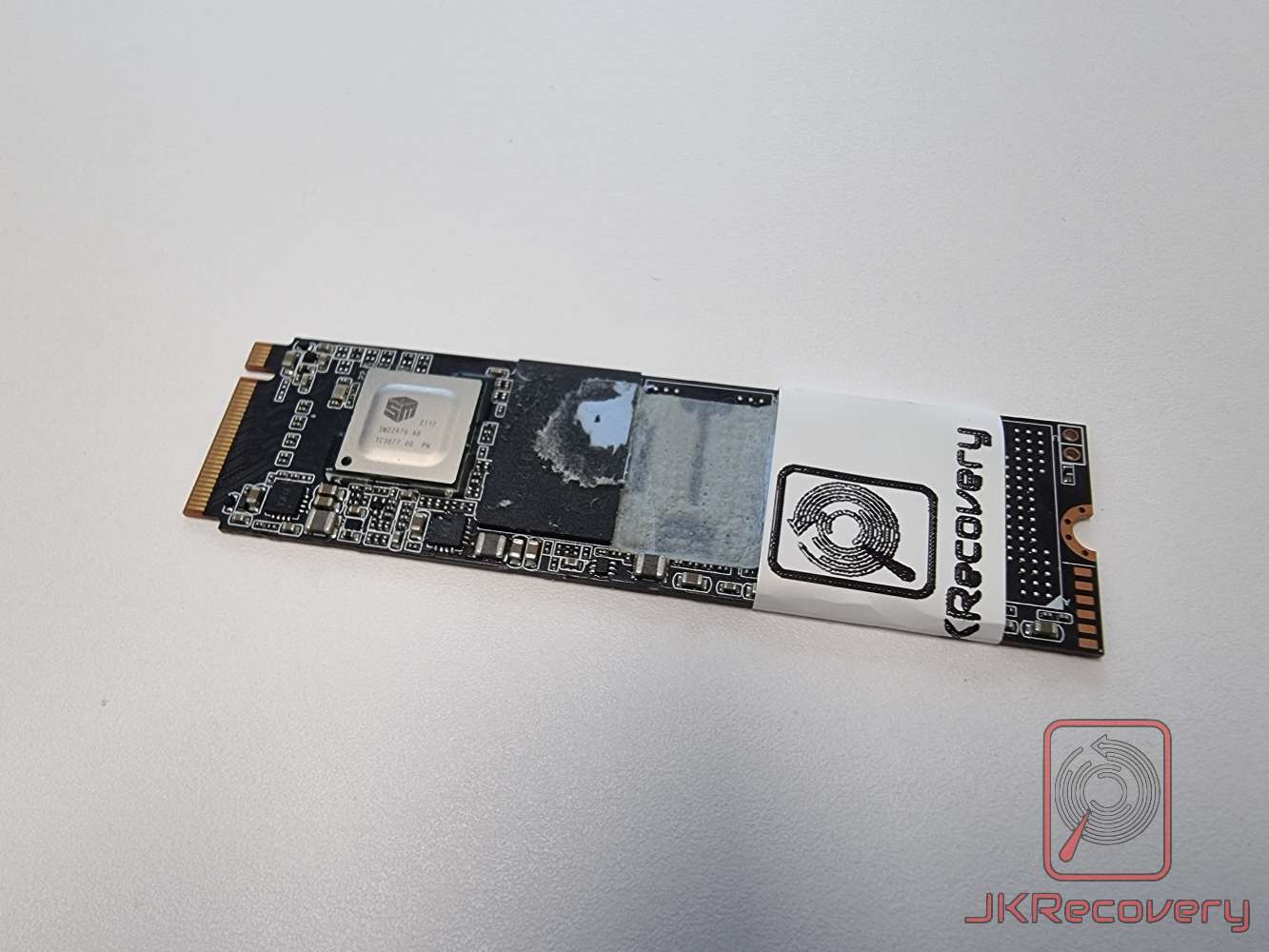 Widok całego dysku SSD M.2 NVMe z kontrolerem Silicon Motion SM2267G z naklejką laboratorium JKRecovery i widocznymi śladami uszkodzeń na powierzchni PCB