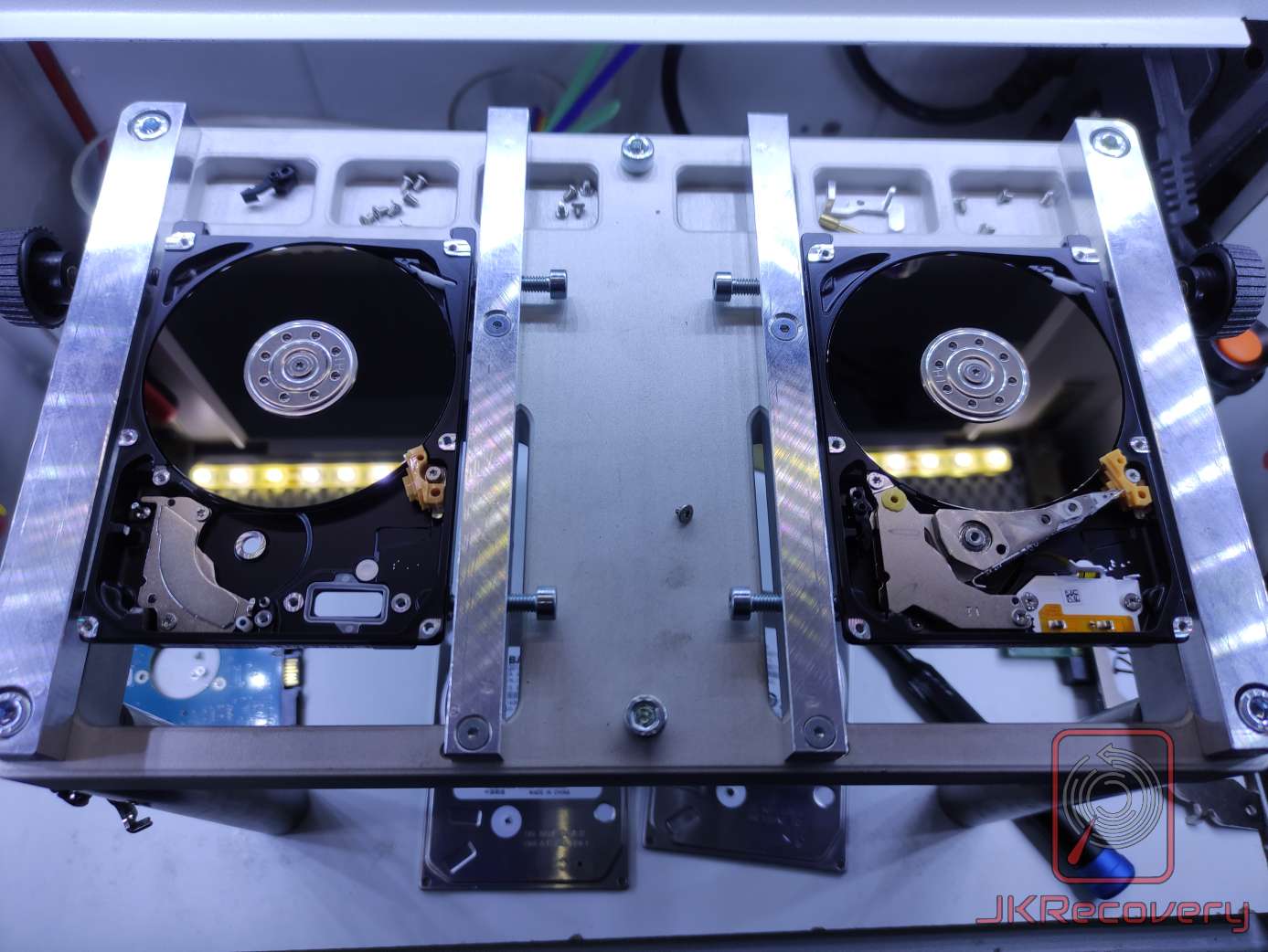 Dwa otwarte dyski twarde HDD zamocowane w specjalistycznym uchwycie laboratoryjnym z oświetleniem LED, z widocznymi talerzami magnetycznymi i głowicami odczytującymi. Zdjęcie wykonane w komorze czystego powietrza podczas procedury odzysku danych.