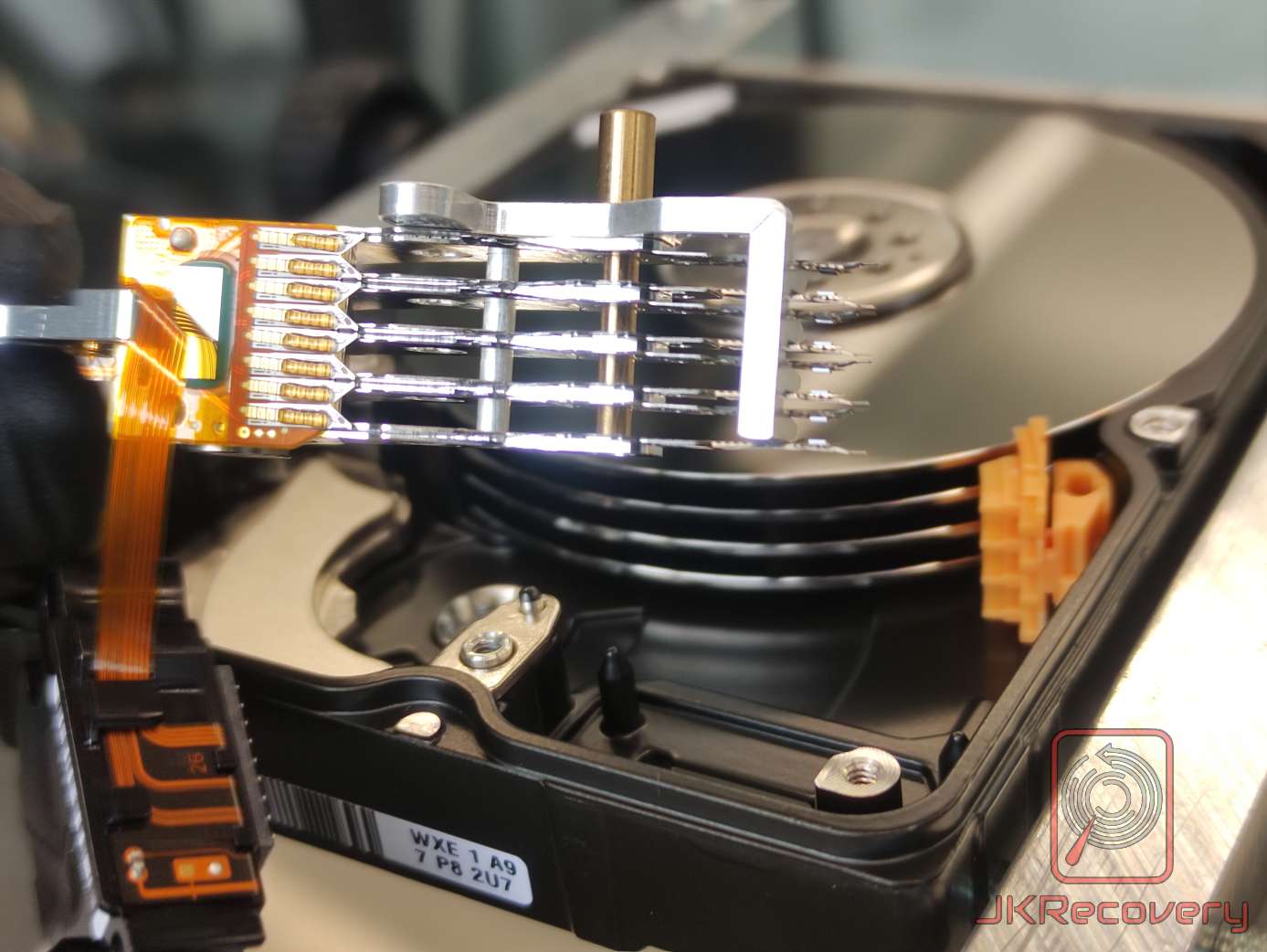 Otwarty dysk twardy HDD z widocznym zespołem głowic odczytująco-zapisujących uniesionych nad wieloma talerzami magnetycznymi, elastyczna taśma FPC podłączona do aktuatora, pomarańczowy złącze cewki głosowej, widok z nieco innego kąta niż zdjęcie pierwsze