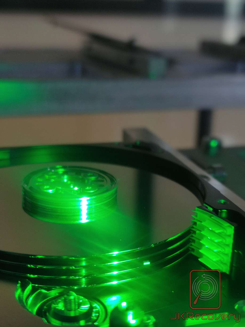 Wnętrze dysku twardego HDD oświetlone zielonym światłem UV z widocznym talerzem, ramieniem głowicy i koncentratorem silnika wrzeciona w laboratorium odzysku danych JKRecovery