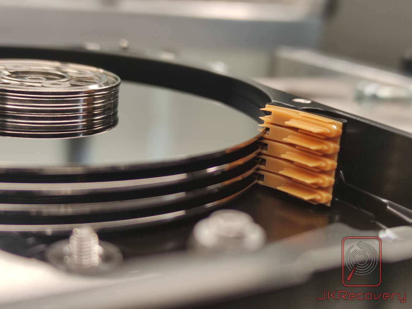 Wnętrze otwartego dysku twardego HDD - widok na stos talerzy magnetycznych z pakietem głowic odczytu/zapisu przyciśniętym do krawędzi talerzy, widoczne uszkodzenie mechaniczne głowic - sklejone ramiona zawieszenia z pomarańczową taśmą flex