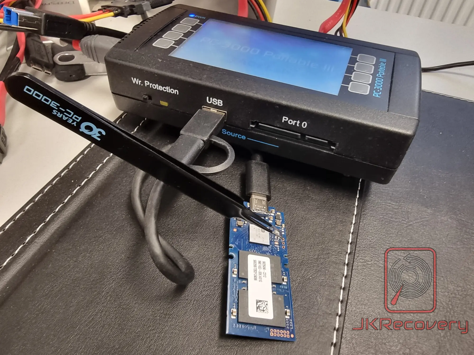 Płytka PCB dysku SSD NVMe podłączona przez USB do urządzenia PC-3000 Portable III firmy ACE Lab, służącego do odzysku danych, na czarnym blacie laboratoryjnym z logo JKRecovery