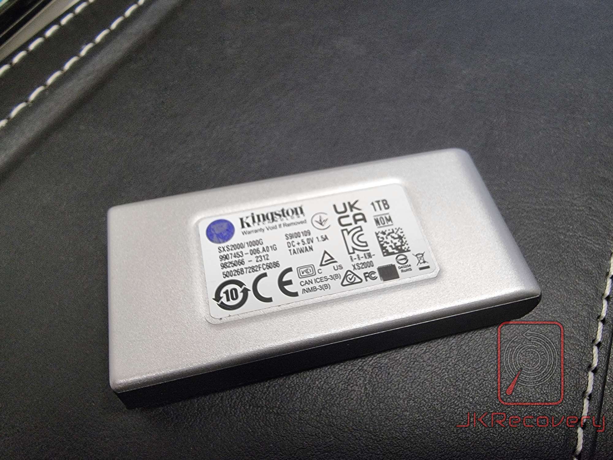 Spód zewnętrznego dysku SSD Kingston XS2000 o pojemności 1TB z widoczną naklejką znamionową zawierającą dane techniczne, numer modelu SXS2000/1000G oraz certyfikaty zgodności